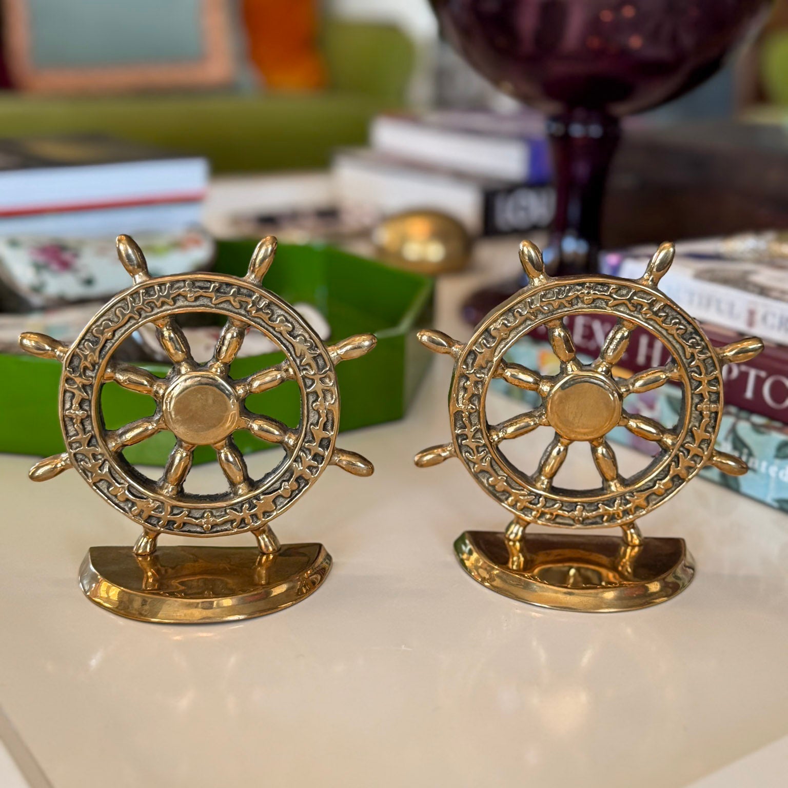 Two gold decorative ship wheels on a surface with books and a glass in the background.