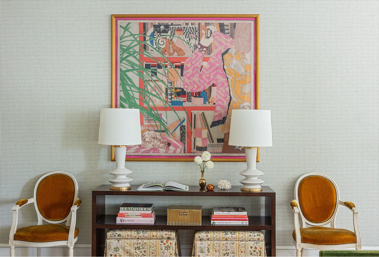 Living room with a colorful abstract painting, two white lamps, a wooden side table with books and a vase, and two orange chairs.